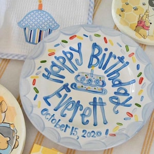 May include: A white ceramic plate with a blue scalloped edge and the words "Happy Birthday Everett" in blue script. The plate features a cake illustration and colorful sprinkles. The date "October 15, 2020" is also written on the plate.
