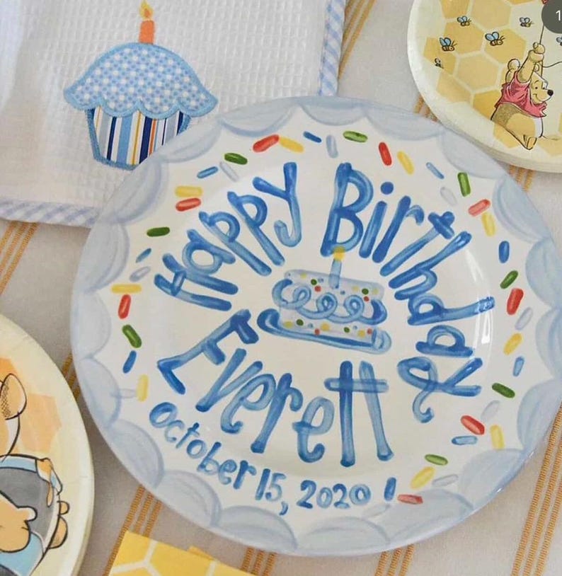 May include: A white ceramic plate with a blue scalloped edge and the words "Happy Birthday Everett" in blue script. The plate features a cake illustration and colorful sprinkles. The date "October 15, 2020" is also written on the plate.