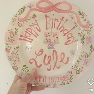 May include: A hand-painted, white ceramic plate with a pink bow and the words "Happy Birthday Lyle" in pink script. The plate features a cake illustration and the date "April 16, 2025". Floral and butterfly accents decorate the rim.