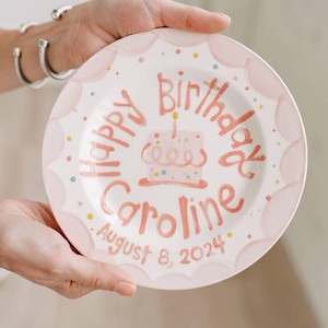 May include: A personalized pink and white plate with the words "Happy Birthday Caroline August 8, 2024" in a handwritten style. The plate features a cake illustration and decorative polka dots.