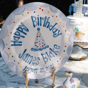 May include: A white ceramic plate with a blue scalloped edge and colorful polka dots. The plate features the words "Happy Birthday James Blake August 21, 2024" in blue script, along with a party hat illustration. The plate is displayed on a gold stand.