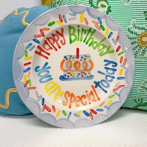 May include: A white ceramic plate with a blue scalloped edge and colorful text that reads "Happy Birthday Today You are Special!" A small cake with a lit candle is in the center, surrounded by colorful sprinkles.