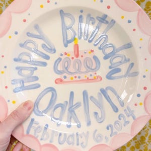 May include: A white ceramic plate with a pink scalloped edge and blue and yellow polka dots. The plate has a blue inscription that says "Happy Birthday Oaklynn February 6, 2024" and a hand-painted blue and pink cake with a yellow candle.