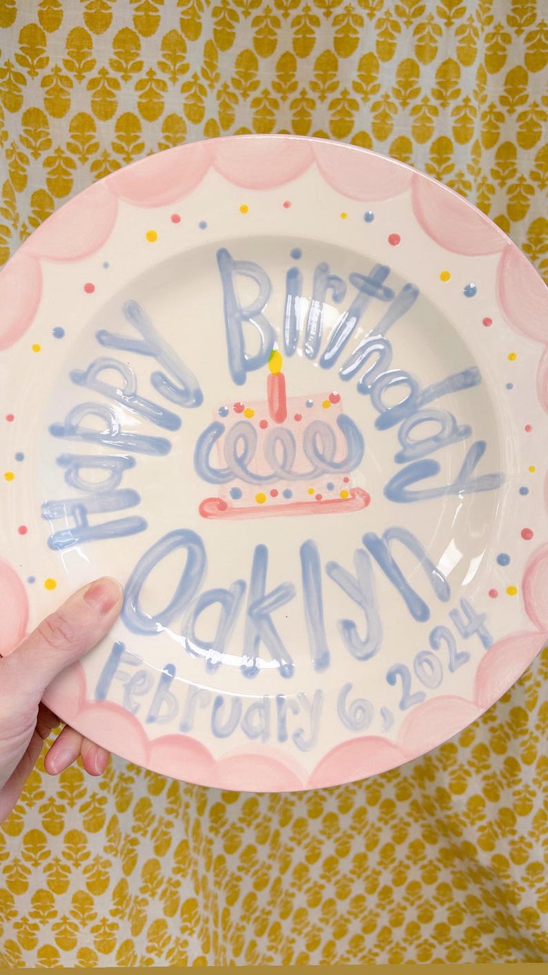 May include: A white ceramic plate with a pink scalloped edge and blue and yellow polka dots. The plate has a blue inscription that says "Happy Birthday Oaklynn February 6, 2024" and a hand-painted blue and pink cake with a yellow candle.
