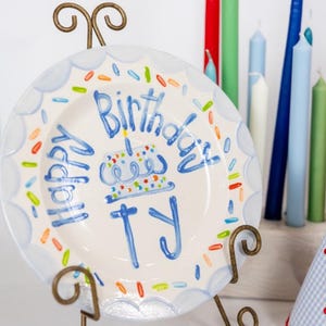 May include: A white ceramic plate with a blue scalloped border and the words "Happy Birthday TY" in blue script. The plate features a cake illustration and colorful sprinkles. It is displayed on a decorative metal stand, with colorful candles in the background.