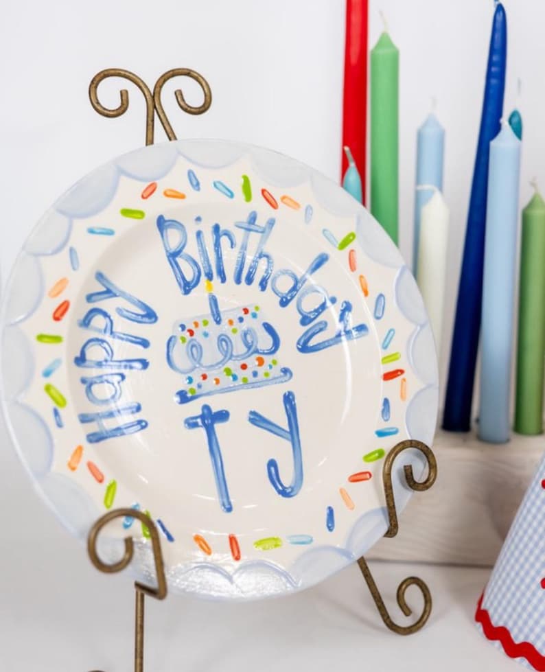 May include: A white ceramic plate with a blue scalloped border and the words "Happy Birthday TY" in blue script. The plate features a cake illustration and colorful sprinkles. It is displayed on a decorative metal stand, with colorful candles in the background.