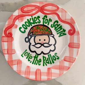 May include: White ceramic plate with a red and white checkered pattern. The plate has a green and red illustration of Santa Claus with the text "Cookies for Santa Love, the Rallos" in green.