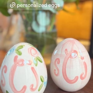 May include: Two white ceramic keepsake eggs with pink initials and floral accents. One egg has blue and green floral details. The text "personalized eggs" is in a white box. The eggs are on a wooden surface.