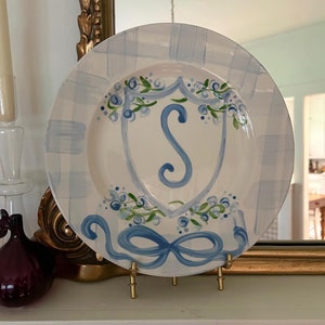 May include: A white ceramic plate with a blue and white plaid pattern. The plate features a hand-painted blue and green floral design with a blue letter "S" in the center. The plate is displayed on a gold metal stand.