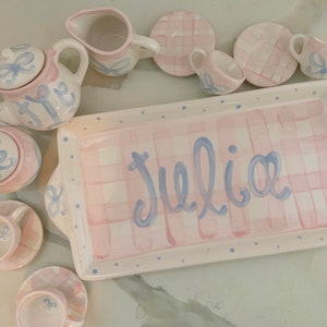 May include: A pink and white checkered serving tray with blue polka dots and the name "Julia" written in blue script. The tray is part of a children's tea set with a teapot, creamer, sugar bowl, and four teacups and saucers.