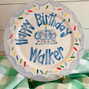 May include: A white ceramic plate with a blue scalloped border and the words "Happy Birthday Walker" in blue. A blue cake with a lit candle is in the center, with colorful sprinkles around the edge. The plate is on a gold stand.