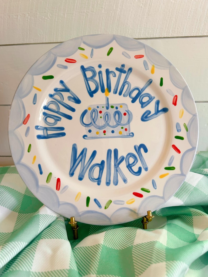 May include: A white ceramic plate with a blue scalloped border and the words "Happy Birthday Walker" in blue. A blue cake with a lit candle is in the center, with colorful sprinkles around the edge. The plate is on a gold stand.