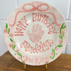 May include: A hand-painted, pink ceramic plate with the words "Happy Birthday Madelyn June 19, 2024" in pink script. The plate features a pink handprint, a pink bow, and green floral accents. The plate is displayed on a gold stand.