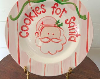 May include: A round, hand-painted ceramic plate with a Santa Claus design. The plate features a pink and white striped border, with the words "Cookies for Santa" in red. A green ribbon design is at the top.