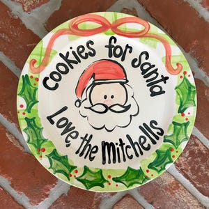 May include: A round, hand-painted ceramic plate with a Christmas theme. The plate features a Santa Claus face in the center, surrounded by the text "cookies for santa" and "Love the Mitchells". The rim has a green and red holly design.