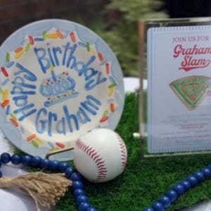 May include: A decorative plate with the words "Happy Birthday Graham" in blue script, a baseball, and a sign that says "Graham Slam". The plate has a blue rim and colorful accents. A blue beaded necklace and a baseball are in the foreground.
