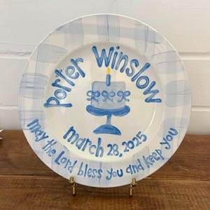 May include: A white ceramic plate with a blue gingham border. The plate features a blue cake illustration and the name "Porter Winslow" in blue script. The date "March 28, 2025" is also written on the plate, along with the phrase "May the Lord bless you and keep you."