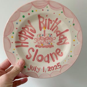 May include: A hand-painted ceramic plate with a pink scalloped border. The plate features the words "Happy Birthday Sloane" in pink script, along with a cake illustration and the date "July 1, 2025". Colorful polka dots decorate the plate.
