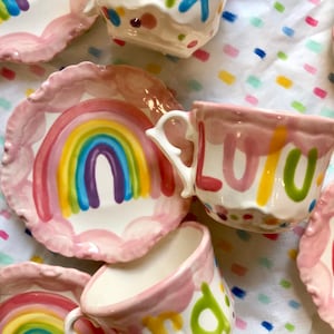 May include: A pink teacup and saucer set with a rainbow design and the word "LULU" written in colorful letters. The saucer has a scalloped edge.