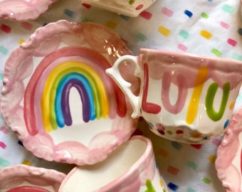May include: A pink teacup and saucer set with a rainbow design and the word "LULU" written in colorful letters. The saucer has a scalloped edge.