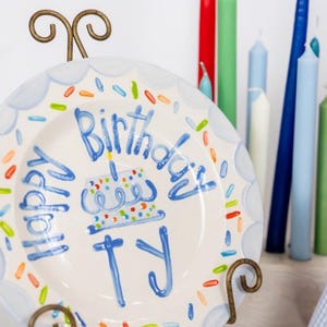 May include: A white ceramic plate with a blue border and colorful sprinkles, held by a decorative metal stand. The plate has the words "Happy Birthday Ty" written in blue, along with a cake illustration. Various colored candles are in the background.