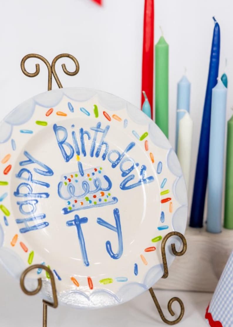 May include: A white ceramic plate with a blue border and colorful sprinkles, held by a decorative metal stand. The plate has the words "Happy Birthday Ty" written in blue, along with a cake illustration. Various colored candles are in the background.
