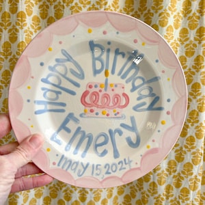 May include: A pink and blue personalized birthday plate with a cake design and the text "Happy Birthday Emery May 15, 2024".