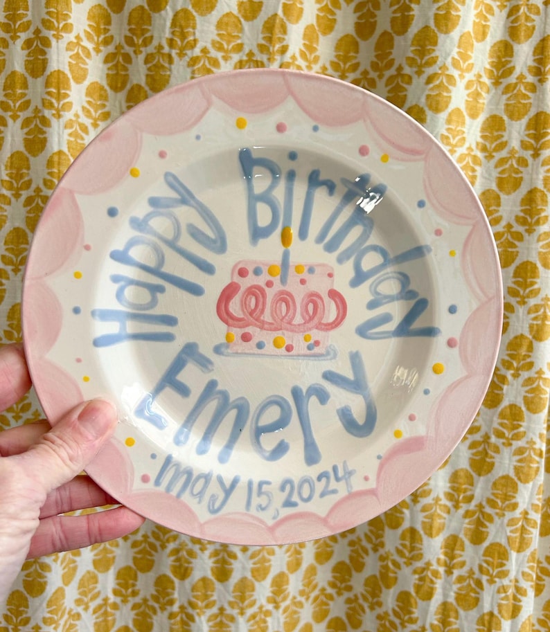 May include: A pink and blue personalized birthday plate with a cake design and the text "Happy Birthday Emery May 15, 2024".