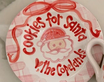 May include: A white ceramic plate with a pink and red plaid pattern. The plate has a red ribbon and the words "Cookies for Santa" written in red. There is a cartoon image of Santa Claus with a pink beard and a red hat. The words "The Copelands" are written in red below Santa.