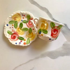 May include: A white teacup and saucer set with a floral design. The design features yellow, red, green, blue, and purple flowers and dots.