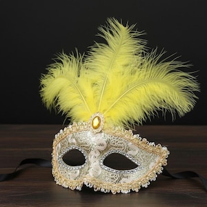 Pode incluir: Uma máscara de baile de máscaras decorativa com um design de renda prateada, acabamento dourado e penas amarelas. A máscara apresenta uma joia central e fitas pretas para fixação. Desenhada para um evento formal ou uma festa à fantasia.