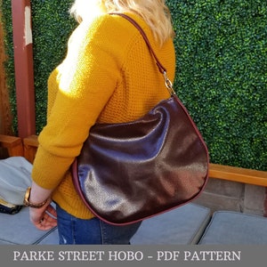 May include: A burgundy leather hobo bag with a brown strap and a gold clasp. The bag is worn by a person wearing a yellow sweater and blue jeans. Parke Street Hobo - PDF Pattern.