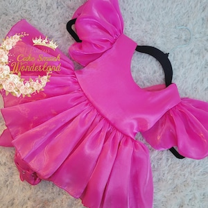 May include: A bright pink, ruffled dress with a matching accessory. The dress features a one-shoulder design with a large, puffy sleeve and a flared skirt. The accessory has the text "Sweet Princess Cake Smash Wonderland" with a crown and floral design.