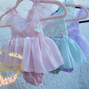 May include: Three pastel-colored baby dresses hanging on pink hangers. The dresses are pink, mint green, and lavender, with tulle skirts and sequin bodices. The dresses are designed for a cake smash or special occasion.