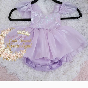 May include: A light purple romper with ruffled sleeves and a layered tulle skirt. The bodice features white floral embroidery and iridescent sequins. A gold logo with the text Sweet Princess Cake Smash Wonderland is visible on the left side.