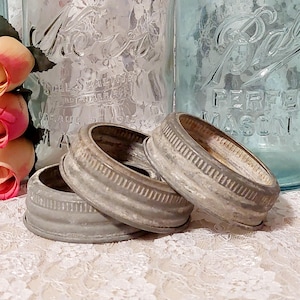 May include: Three vintage, weathered metal jar lids are stacked on a lace surface. The lids are silver-toned with ribbed edges, suggesting a rustic or antique aesthetic. In the background, there are glass jars and pink roses.