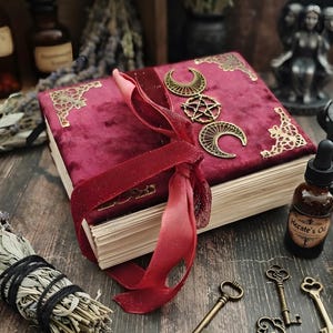 Peut inclure: Un livre rectangulaire relié en velours rouge profond, orné de coins en métal doré et d'un emblème central représentant une triple lune et un pentagramme. Le livre est fermé par un ruban de velours rouge et possède des pages en papier crème.