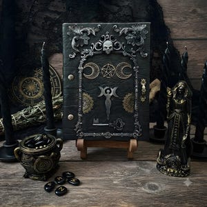 Peut inclure: Un journal relié en cuir noir orné d'un cadre argenté avec un crâne au sommet. La couverture présente un symbole central de triple lune, un pentagramme, une silhouette féminine stylisée et une clé décorative, le tout en métal argenté et doré.