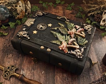Puede incluir: Un libro rectangular negro con cubierta texturizada que presenta un emblema central circular de árbol, dos estrellas pequeñas y un grupo de setas y hojas de helecho tridimensionales. Las esquinas tienen adornos de filigrana metálica.