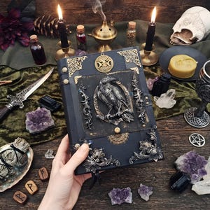 May include: A black hardcover book featuring a central silver-toned raven relief, ornate metal corner accents, and a circular gold-toned triple moon emblem on the cover. The book is bound with dark material and includes intricate decorative metal filigree.