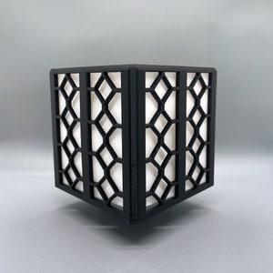May include: A black, geometric-patterned cube-shaped decorative lamp. The lamp features a lattice design on its sides, with a white interior that diffuses light. The lamp is designed to sit on a flat surface.