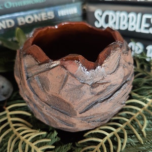 May include: A small, brown, ceramic planter with a textured, rough surface. The planter has a wide, rounded opening and a slightly uneven rim.