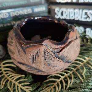 May include: A small, handmade ceramic bowl with a dark brown glaze and a textured, rough surface. The bowl has a wide, rounded opening and a deep interior.