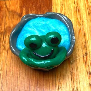 Könnte beinhalten: Eine handgefertigte grüne Froschfigur mit großen Augen mit schwarzen Pupillen und einem lächelnden Mund. Der Frosch sitzt in einem blauen Pool in einer grauen Schale. Die Figur steht auf einer Holzoberfläche.