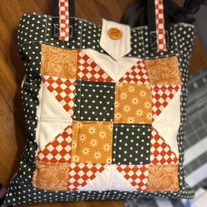 May include: A handmade tote bag with a patchwork design. The bag features a star pattern in the center, with orange, white, and dark green fabrics. The bag has black straps with red and white checkered accents and a wooden button closure.