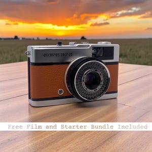 Op de afbeelding: Olympus Trip 35 camera met een zilveren bovenkant, bruine lederlook behuizing en zwarte lens. De camera staat op een houten oppervlak met een zonsondergang op de achtergrond. De tekst "Olympus Trip 35" is zichtbaar.