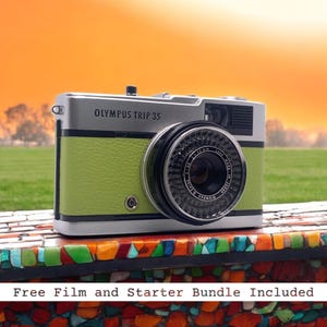 May include: A green and silver Olympus Trip 35 film camera with a black lens. The camera is sitting on a colorful mosaic surface.