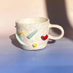 Puede incluir: Taza de cerámica blanca con una superficie texturizada y formas geométricas de colores. La taza tiene un asa y está decorada con detalles rojos, amarillos y azules.