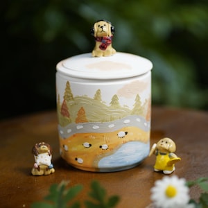 Puede incluir: Un tarro de cerámica blanco con tapa, con un paisaje pintado a mano con árboles, una carretera y ovejas. Una pequeña figura de perro con orejeras y bufanda se encuentra sobre la tapa. Dos figuras de perro adicionales están cerca del tarro.