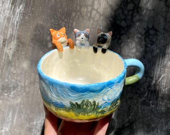 May include: A ceramic mug with a blue and white landscape design, featuring three small cat figurines perched on the rim. The mug has a light blue handle and a cream-colored interior.